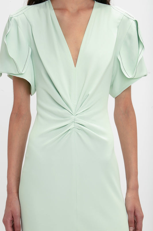 Close-up of a woman wearing a mint green Gathered V-Neck Midi Dress in Jade by Victoria Beckham with short ruffled sleeves and a waist-defining pleat.