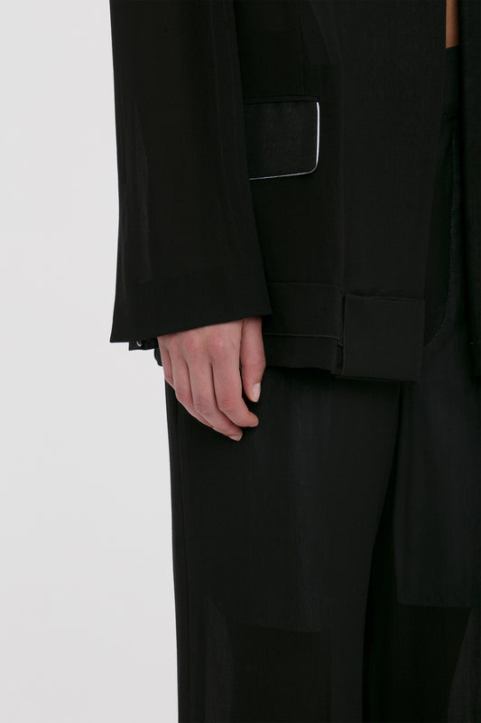 Close-up of a person wearing an all-black outfit, focusing on the hand and lower part of the sleeve, showcasing the contemporary flair of a Fold Detail Tailored Jacket In Black by Victoria Beckham.