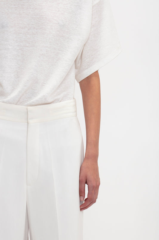 Close-up of a person wearing a white t-shirt and contemporary cool Waistband Detail Straight Leg Trouser In White by Victoria Beckham, showing their torso and part of their right arm against a plain background.