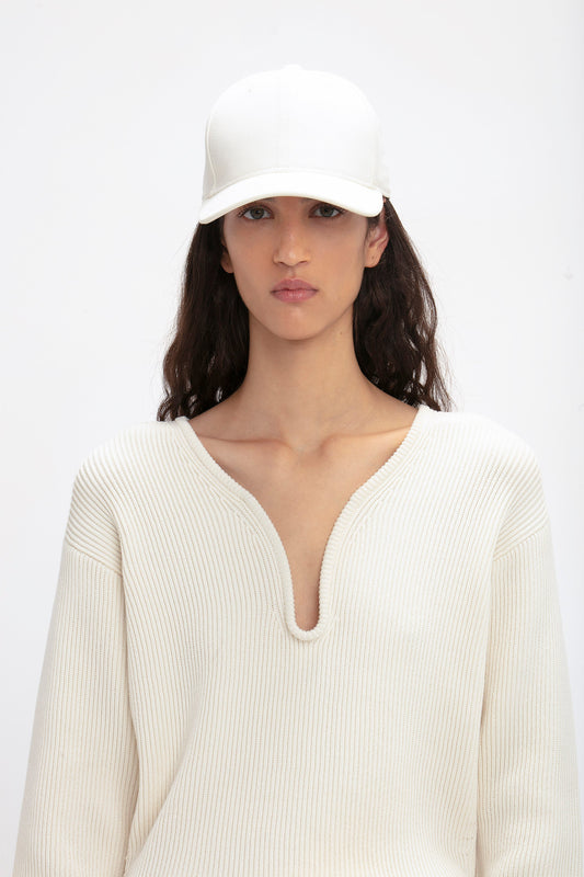 A woman wearing a white Logo Cap in Antique White by Victoria Beckham and cream ribbed sweater, looking directly at the camera with a neutral expression.
