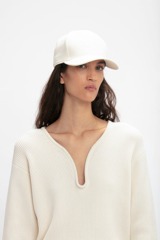 Woman in a white Victoria Beckham Logo Cap In Antique White and ribbed sweater, looking at the camera with a neutral expression against a plain background.