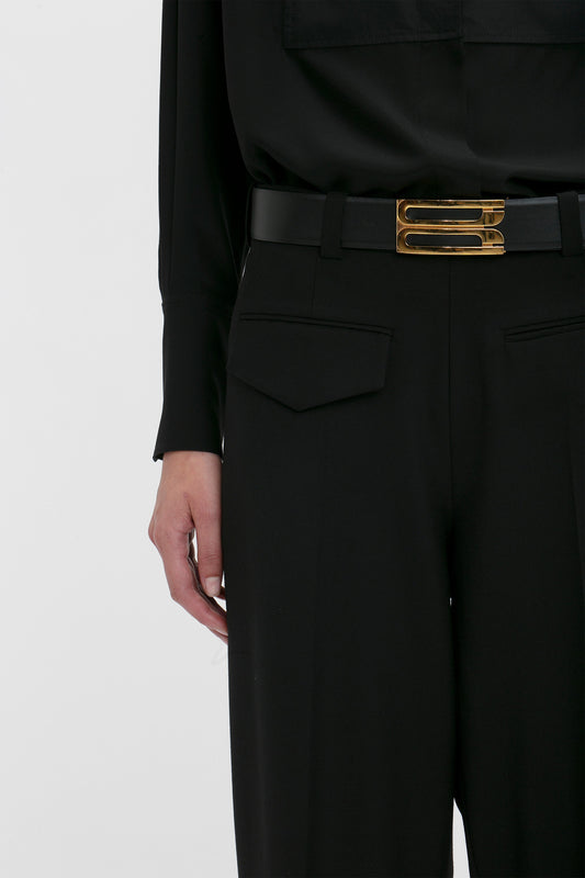 A modern woman dons Victoria Beckham's Reverse Front Trouser In Black with a gold-buckled black belt, paired with a long-sleeve black shirt. The image highlights the contemporary silhouette, focusing on the waist and part of the shirt.