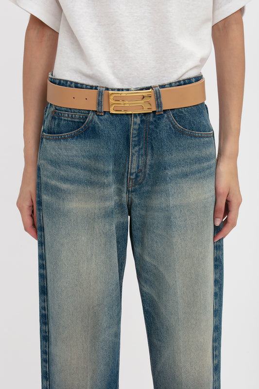 Close-up of a person wearing blue jeans and a brown Victoria Beckham Jumbo Frame Belt In Camel Leather, featuring gold hardware, hands casually placed in pockets.