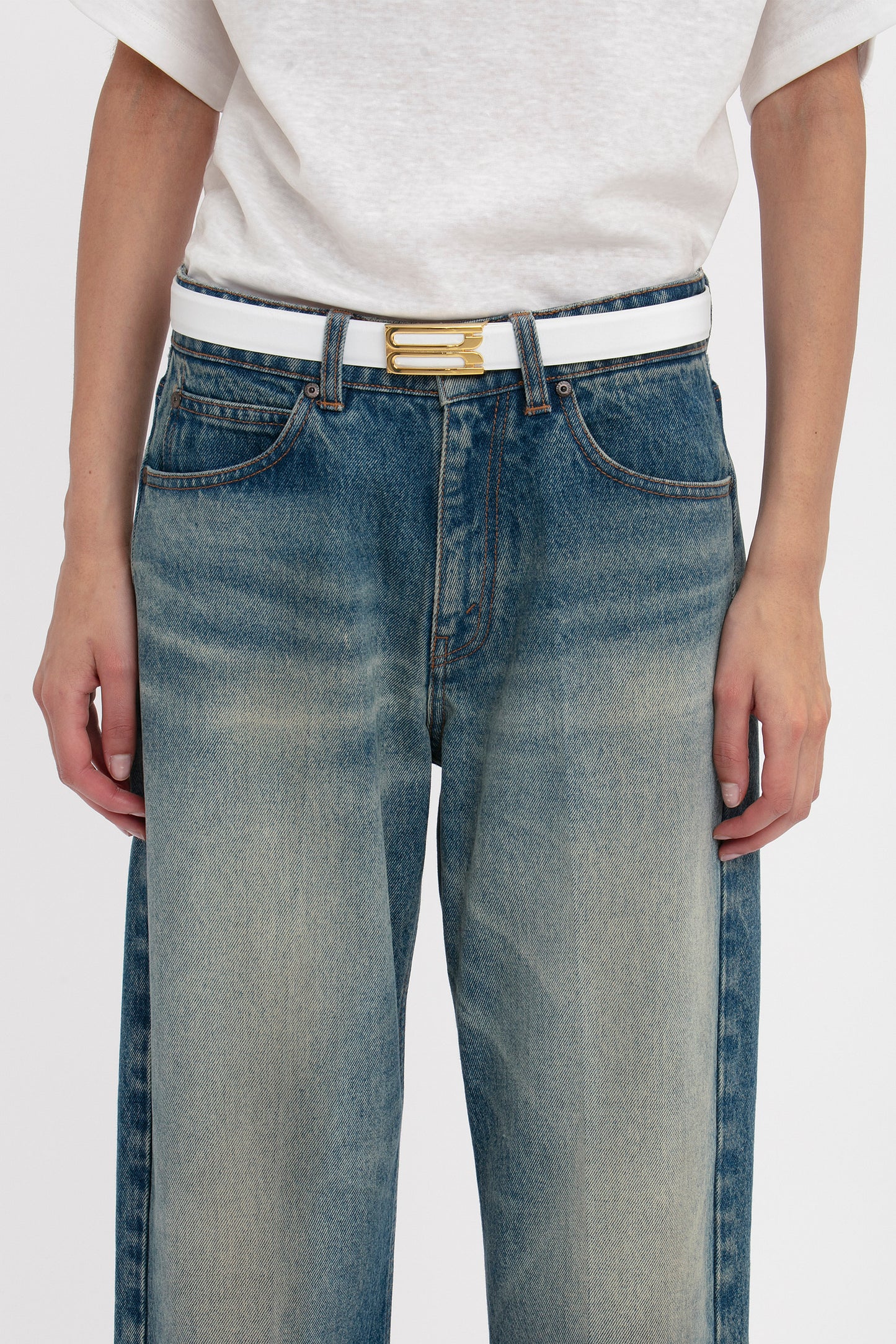 A person wearing a white t-shirt tucked into faded blue jeans with a Victoria Beckham Frame Belt In White Leather featuring gold hardware, showing hands resting at their sides.