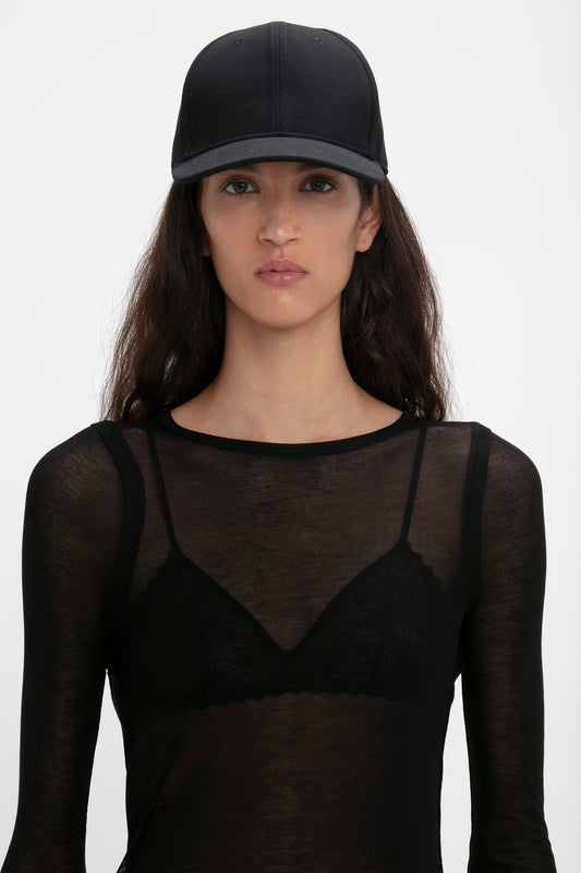 A woman with long brown hair wearing a sheer black top under a black Exclusive Logo Cap In Black by Victoria Beckham, looking directly at the camera.