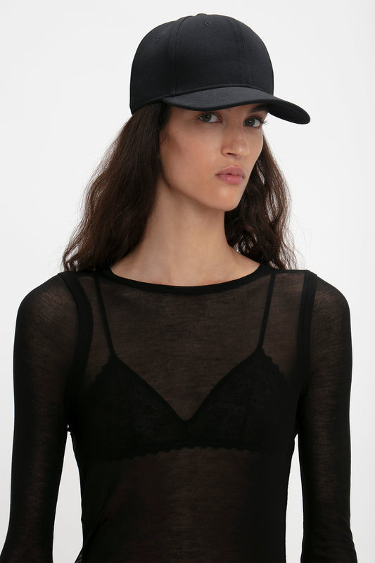 Woman wearing a Victoria Beckham Exclusive Logo Cap in Black and sheer black top with lace details, looking to the side against a white background.