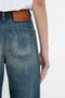 Rear view of a person wearing blue Victoria Beckham Alina High Waisted Jean In Indigrey Wash with seventies-style pockets and a brown leather label, paired with a white T-shirt tucked in. The person's arm is visible, resting by their side.