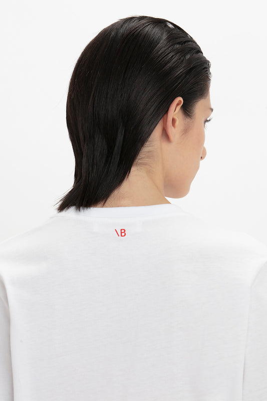 Rear view of a person with shoulder-length dark hair, wearing a Victoria Beckham organic cotton white t-shirt with a small red logo on the collar.