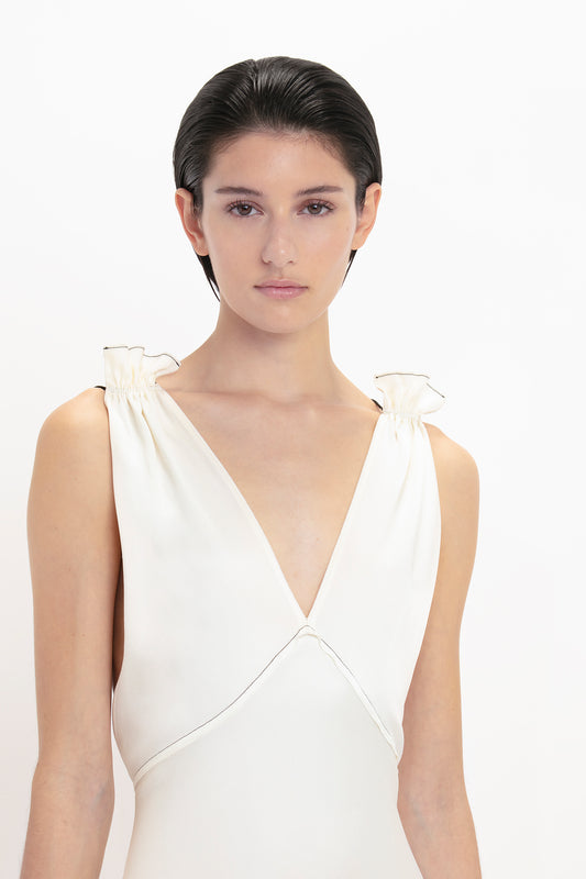 A person with short hair is wearing a sleeveless white Gathered Shoulder Floor-Length Cami Gown In Ivory by Victoria Beckham, made from elegant crepe back satin, standing against a plain white background.