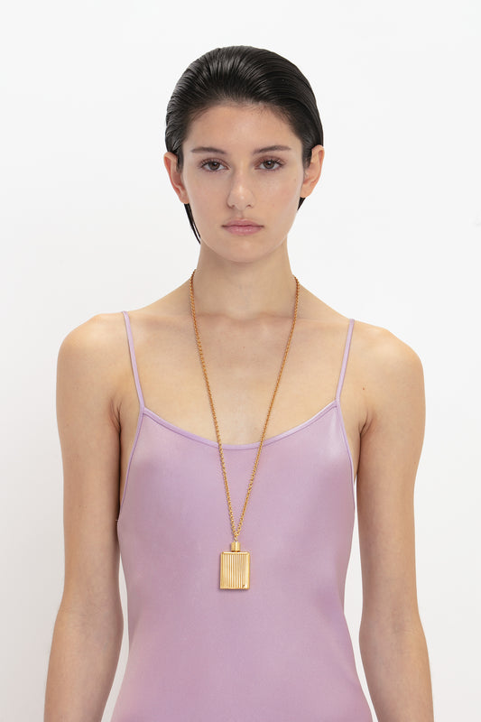 A woman with slicked-back hair wearing a lavender Victoria Beckham camisole slip dress with a deep V neckline and a gold pendant necklace, standing against a white background.