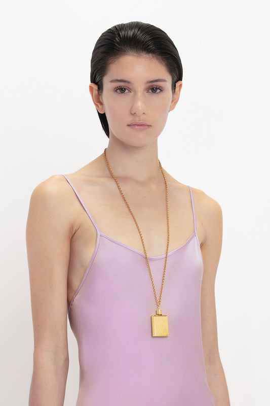 A woman in a Victoria Beckham Low Back Cami Floor-Length Dress In Rosa, wearing a long gold necklace with a rectangular pendant, standing against a white background.