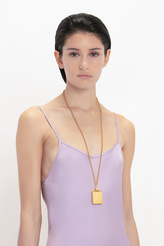 Woman wearing a Victoria Beckham Low Back Cami Floor-Length Dress In Petunia and a long gold perfume bottle necklace, standing against a white background.