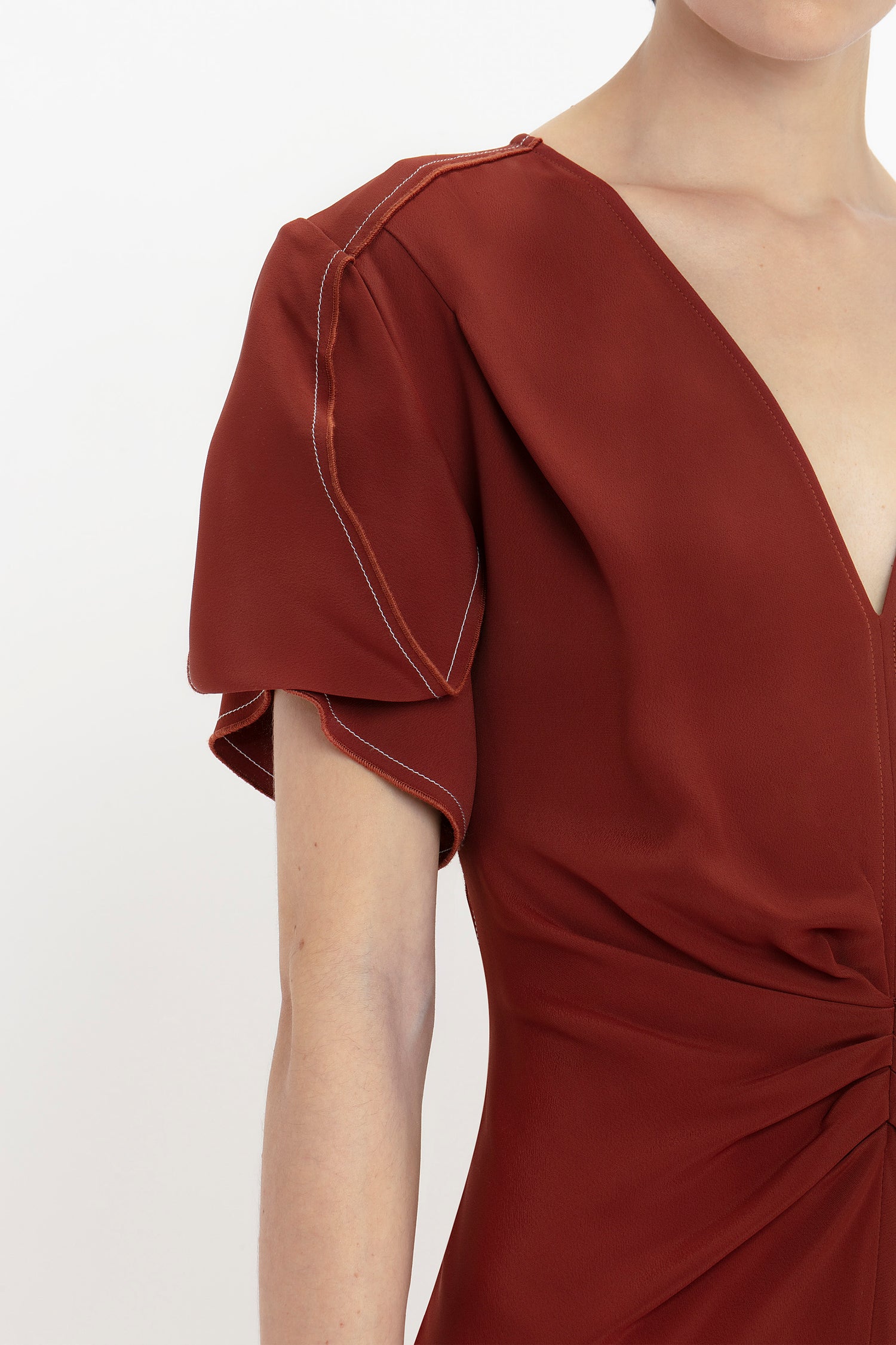 A person is wearing a deep red Gathered V-Neck Midi Dress In Russet by Victoria Beckham with short, puffed sleeves and visible white stitching along the sleeve seams. The figure-flattering stretch fabric showcases a waist-defining pleat detail that adds elegance to the look.