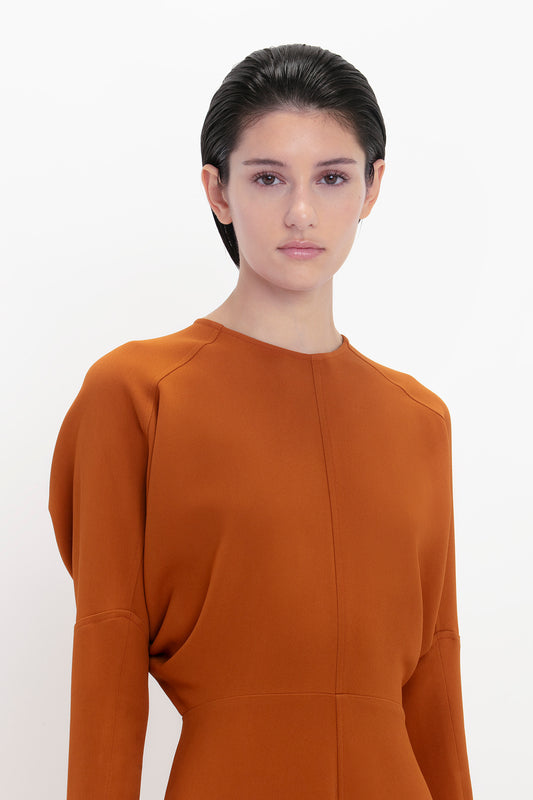 Woman with dark hair, wearing a Victoria Beckham dolman midi dress in russet, standing against a white background, looking directly at the camera.