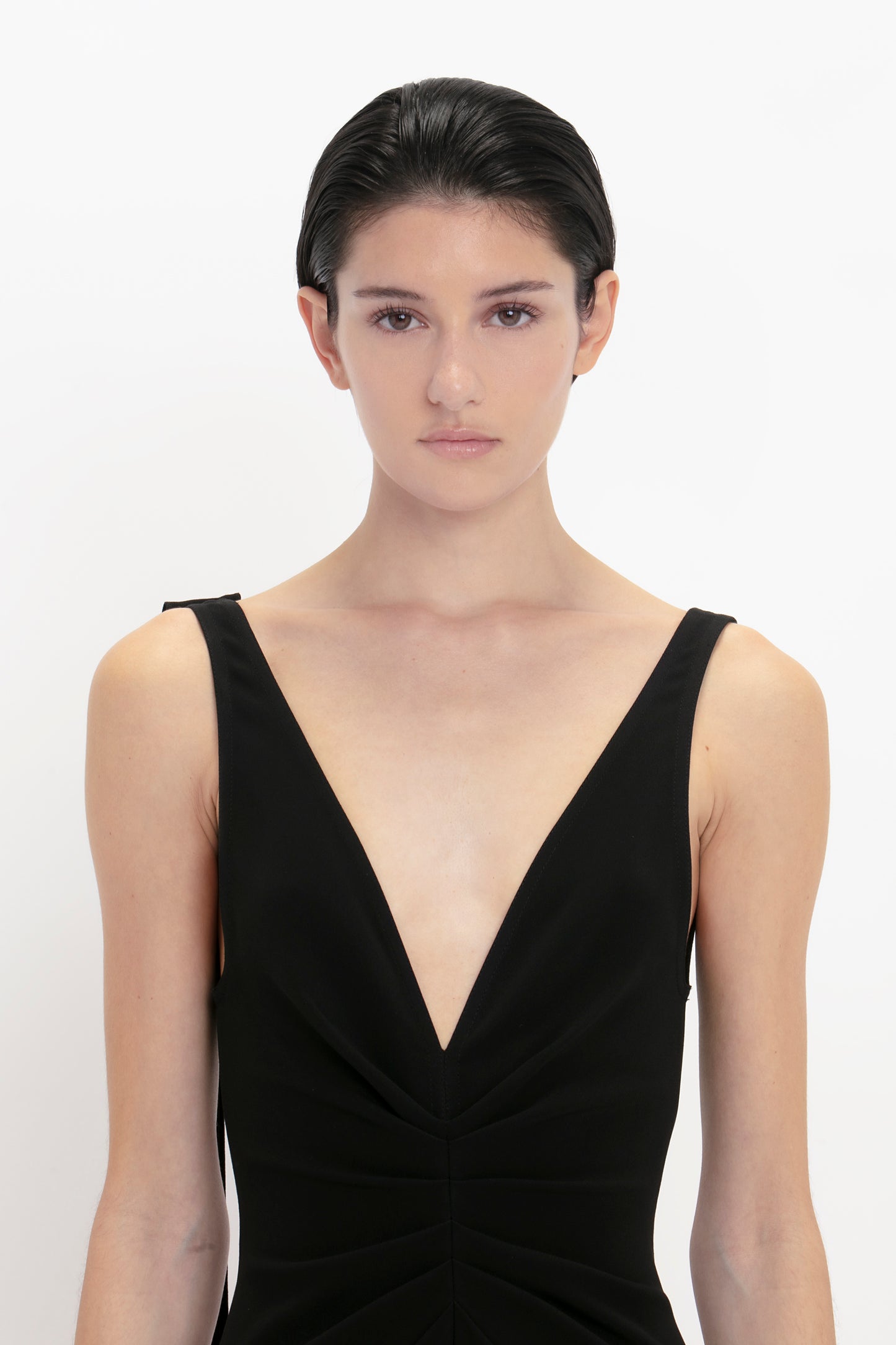 A person with short dark hair is wearing a sleeveless black V-Neck Gathered Waist Floor-Length Gown In Black by Victoria Beckham. Their expression is neutral, and they are standing against a plain white background.