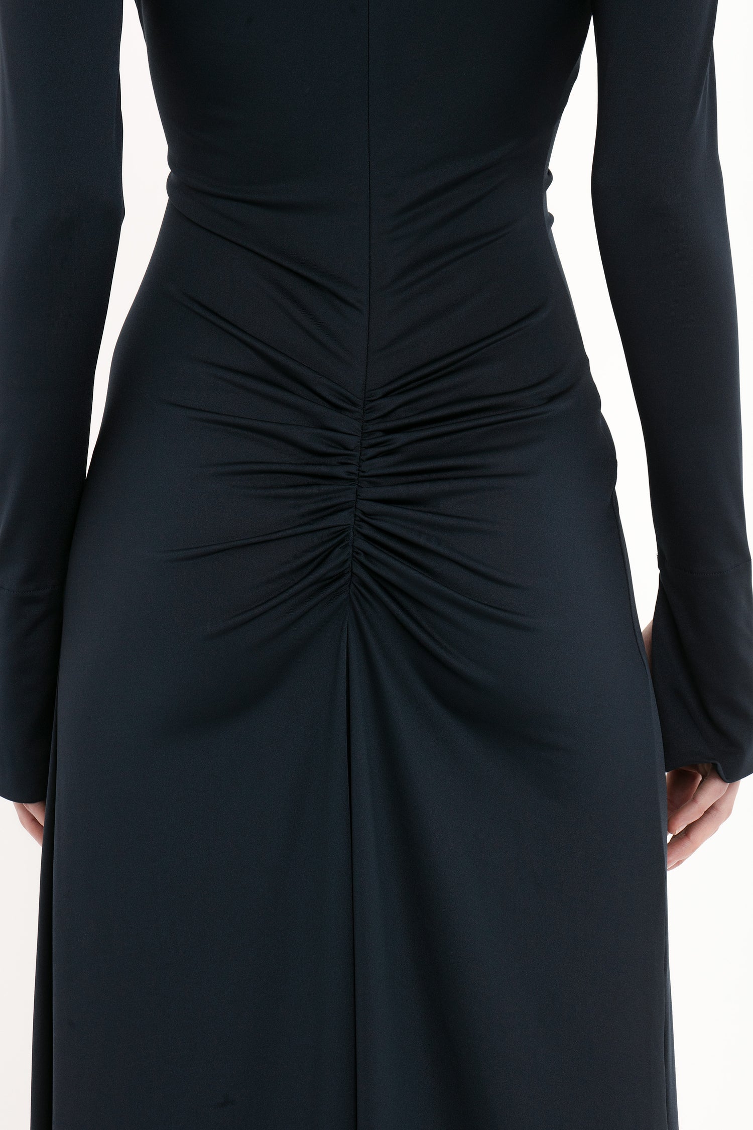 Back view of a person wearing a fitted, long-sleeve black Ruched Detail Floor-Length Gown In Midnight with sophisticated ruching at the lower back, reminiscent of the timeless elegance seen in Victoria Beckham's evening gowns.