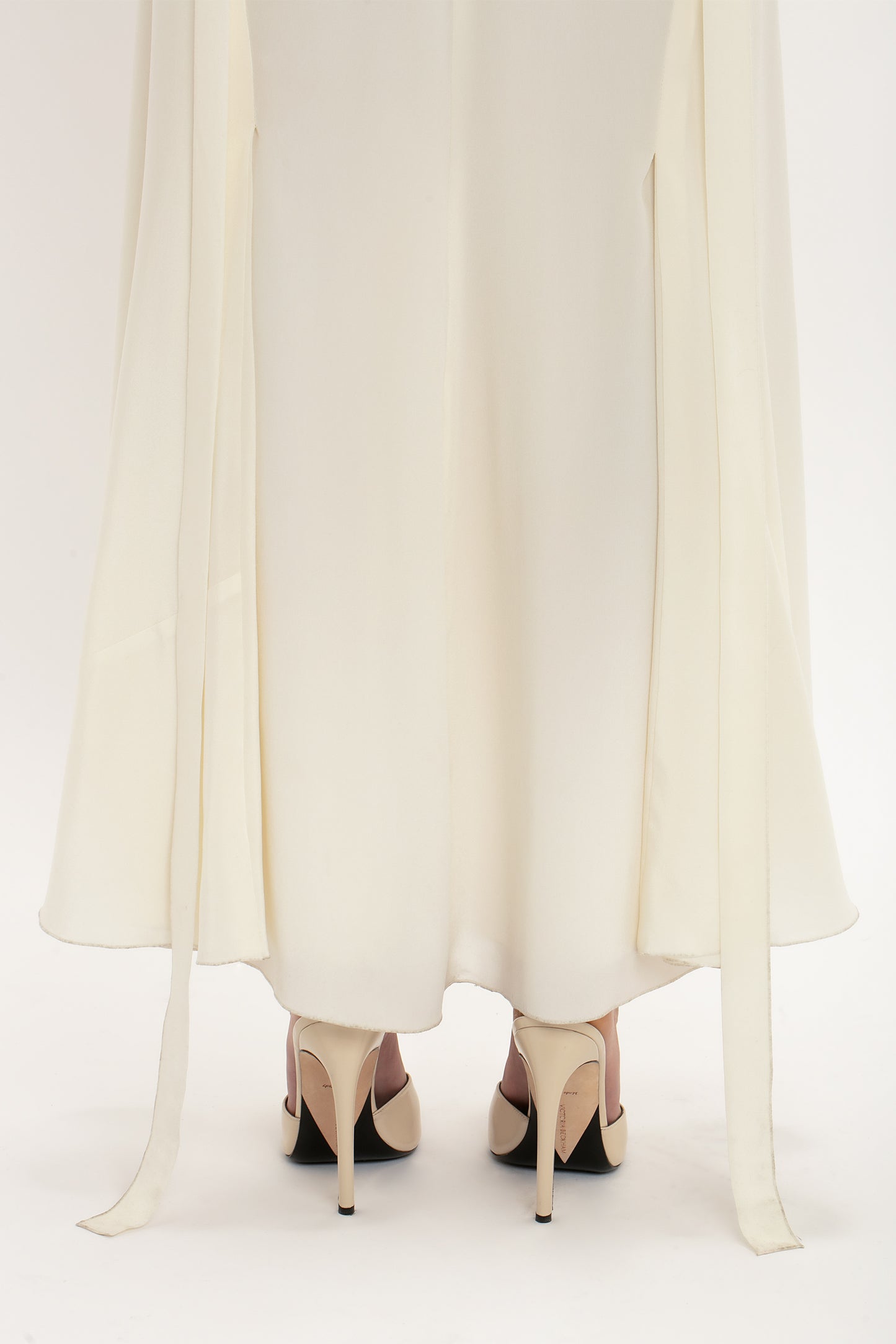 A person wearing an elegant white Ruffle Detail Midi Dress In Ivory by Victoria Beckham and beige high heels stands on a white background. Two long fabric strips hang from the sleeves of the dress.