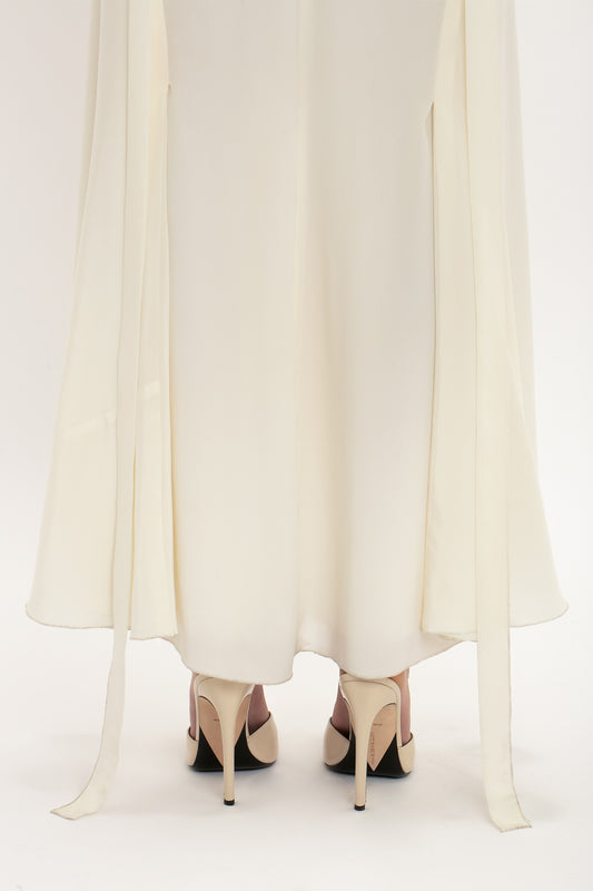 A person wearing an elegant white Ruffle Detail Midi Dress In Ivory by Victoria Beckham and beige high heels stands on a white background. Two long fabric strips hang from the sleeves of the dress.
