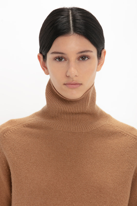 Woman with dark hair in a brown lambswool Victoria Beckham polo neck jumper looking at the camera, neutral expression, white background.
