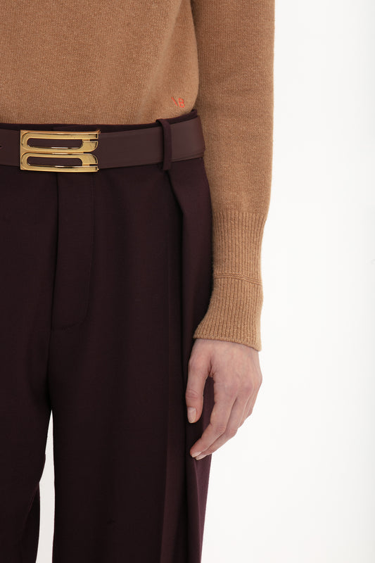 Close-up of a person wearing a Victoria Beckham lambswool polo neck jumper in tobacco and maroon trousers with a gold buckle belt, focusing on the midsection.