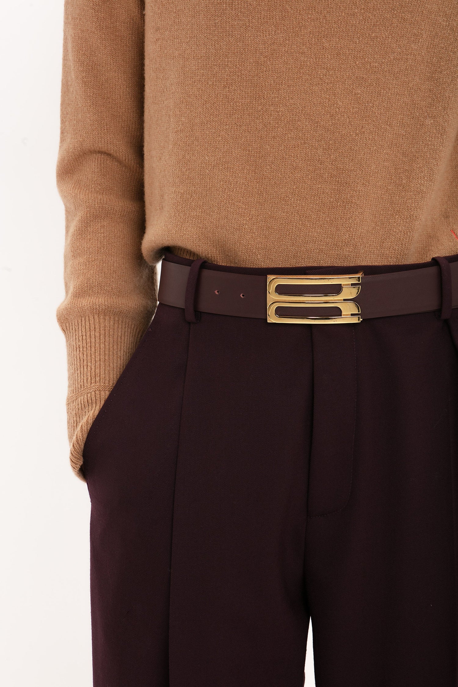 Close-up of a person wearing a tan sweater, Victoria Beckham Asymmetric Chino Trouser In Deep Mahogany with a narrow leg silhouette, and a maroon belt with a decorative gold buckle. Hands are in the pockets.