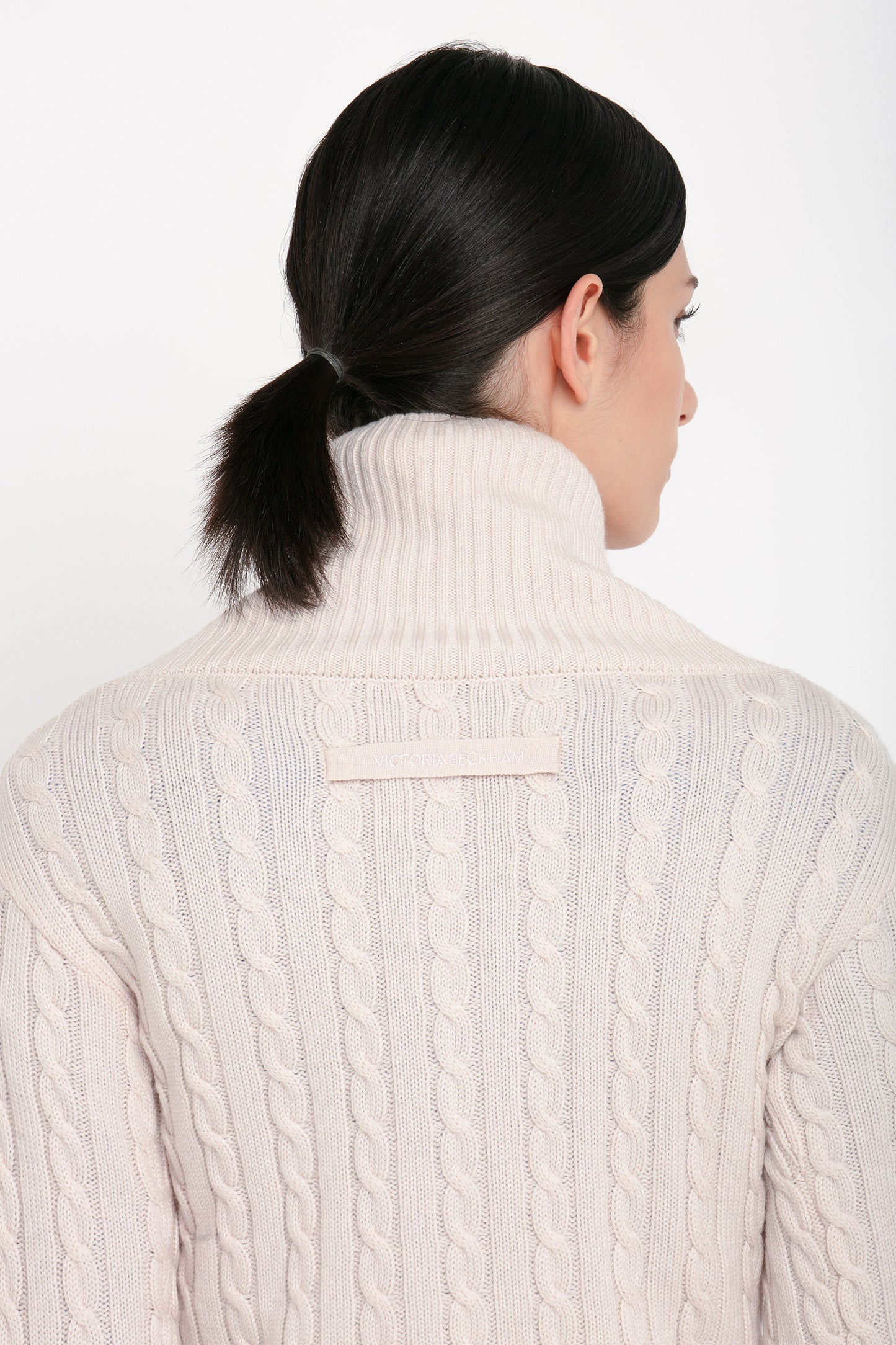 A person with dark brown hair tied in a low ponytail is wearing a Victoria Beckham Wrap Detail Jumper In Bone made from cozy cabled merino wool, seen from behind.