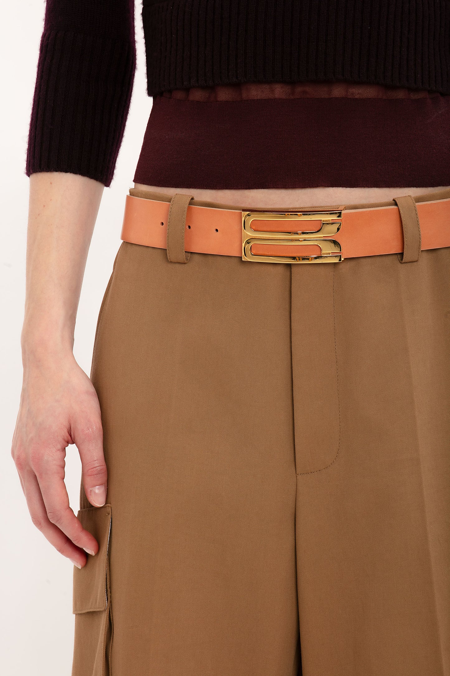 A person wearing tan high-waisted pants with a double buckle tan belt and a dark cropped Victoria Beckham Wrap Detail Jumper In Brown, exuding an air of Victoria Beckham-inspired sophistication.
