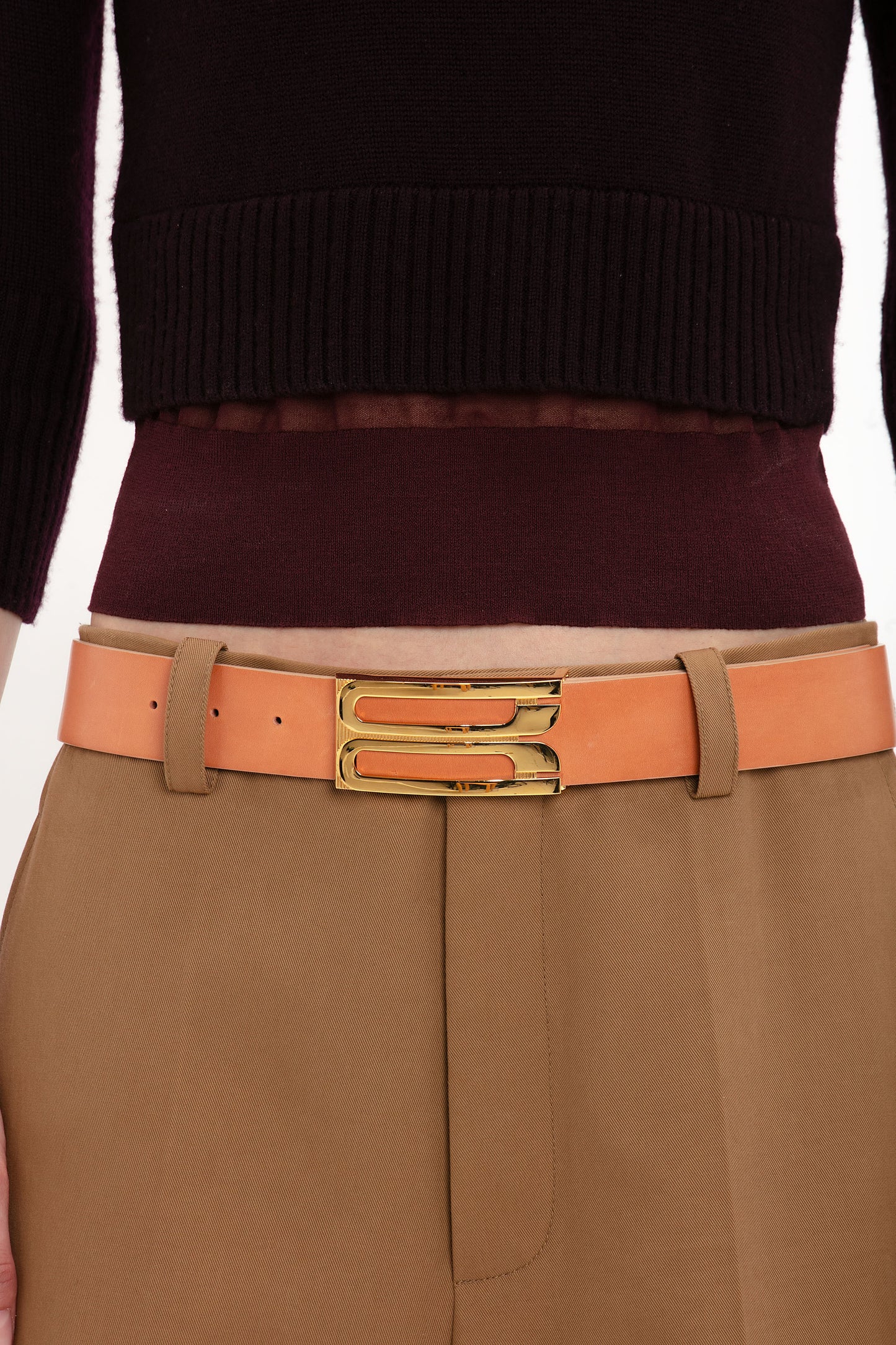 Close-up of a person wearing a tan belt with a gold buckle over high-waisted brown trousers and a dark maroon Victoria Beckham Wrap Detail Jumper In Brown.
