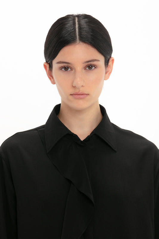 Young woman with dark hair wearing a Victoria Beckham Asymmetric Ruffle Blouse In Black, standing against a white background, looking directly at the camera.