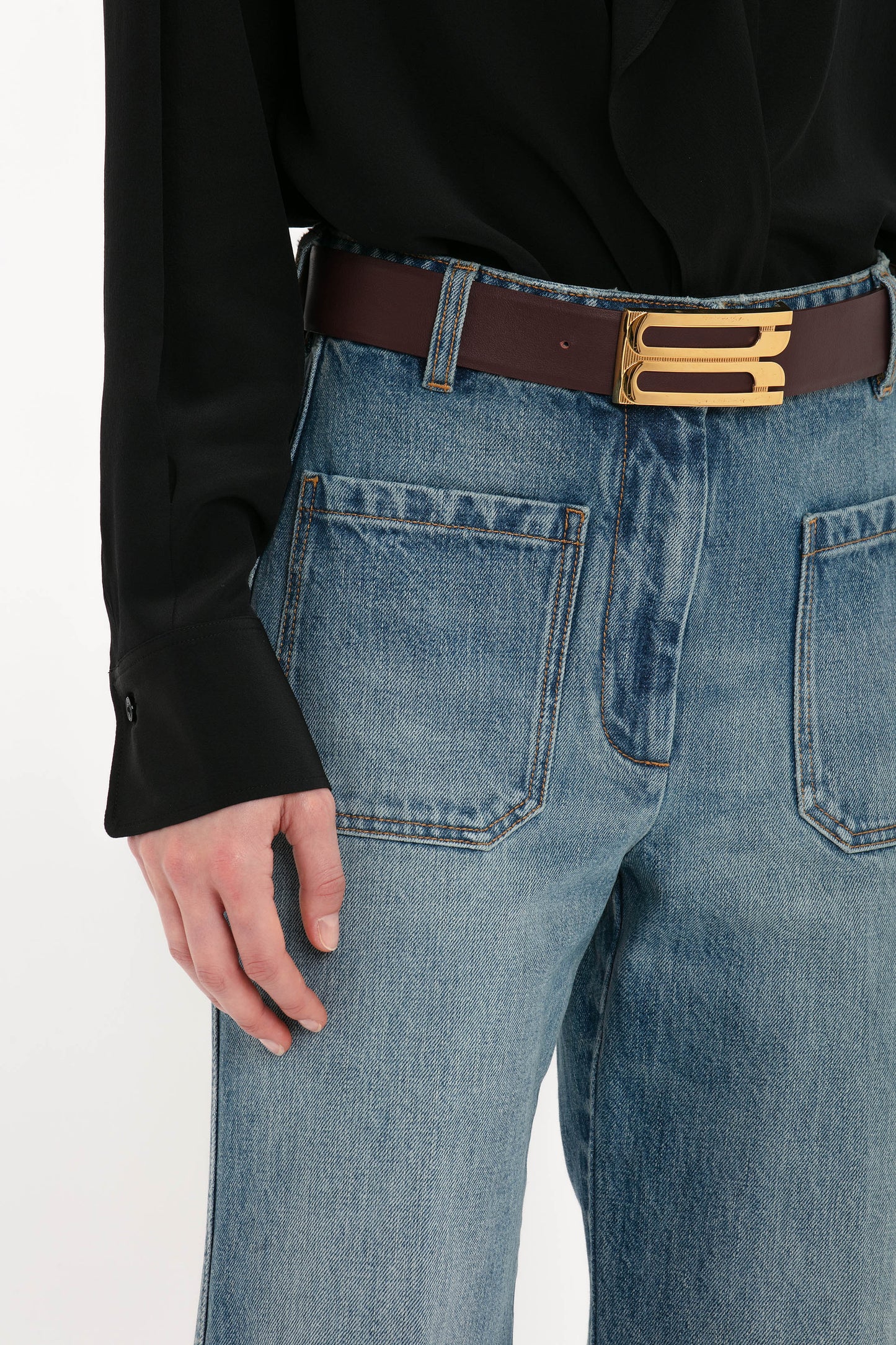 A person wearing Victoria Beckham Alina High Waisted Jean In Worn Blue Wash with seventies-inspired patch pockets, complemented by a black shirt and a brown belt featuring a gold buckle.