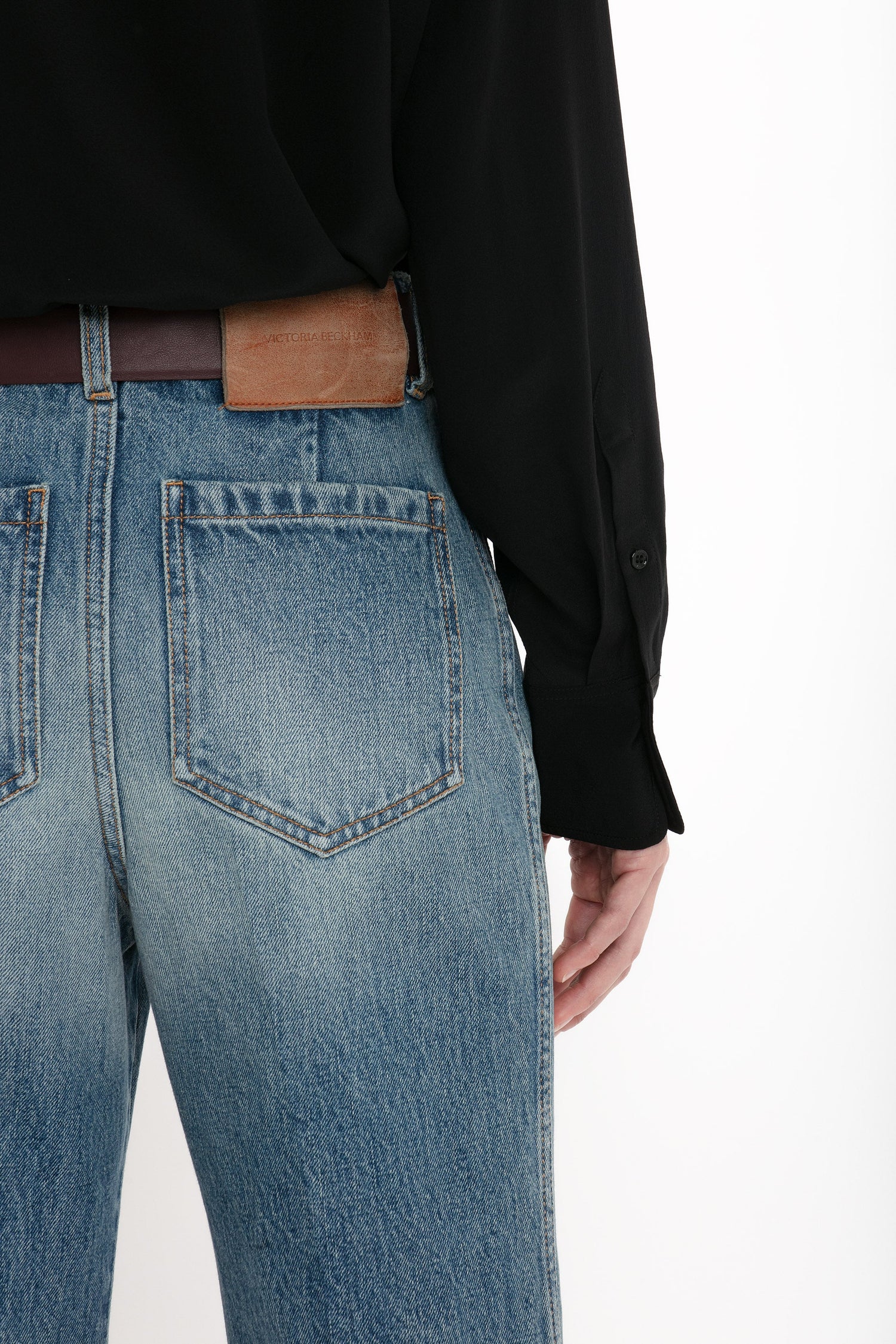 A person wearing a black shirt is shown from the back, displaying a pair of Victoria Beckham Alina High Waisted Jeans In Worn Blue Wash with seventies-inspired patch pockets and a brown leather patch above the right back pocket.