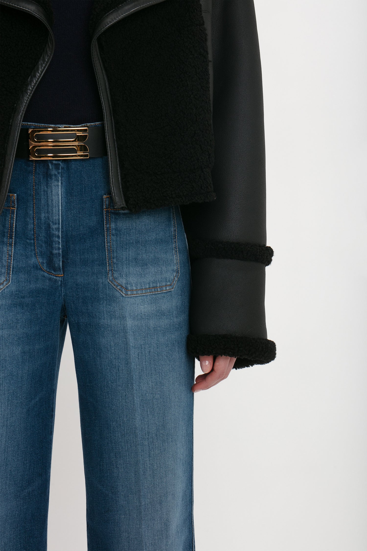 Person wearing a Victoria Beckham Shearling Jacket In Black, dark top, and blue high-waisted jeans with front pockets and a gold belt.