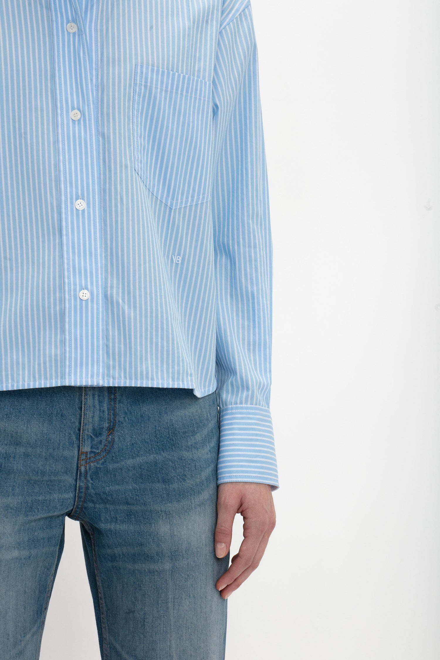 A person wearing a Cropped Long Sleeve Shirt In Marina-White by Victoria Beckham and blue jeans, standing against a plain white background. The photo is cropped at the neck and thighs.