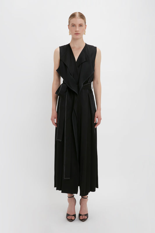 A person stands against a plain white background, donning the Trench Dress In Black by Victoria Beckham with matching wide-leg pants. The look, part of a chic black colourway, is accessorized with black open-toe heels and small earrings.
