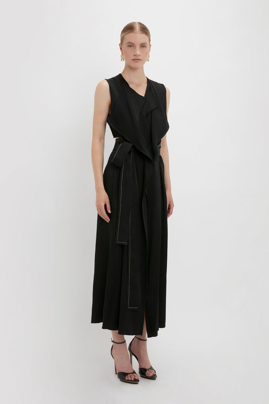 A woman stands in a Trench Dress In Black by Victoria Beckham with a belted waist and black high-heeled sandals against a plain white background, embodying the essence of utilitarian outerwear.