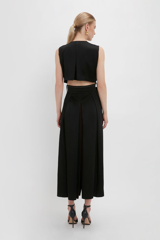 A person with blonde hair, wearing a black sleeveless top and black wide-leg pants, stands facing away, showcasing the back details of the Victoria Beckham Trench Dress In Black in a sleek black colourway.