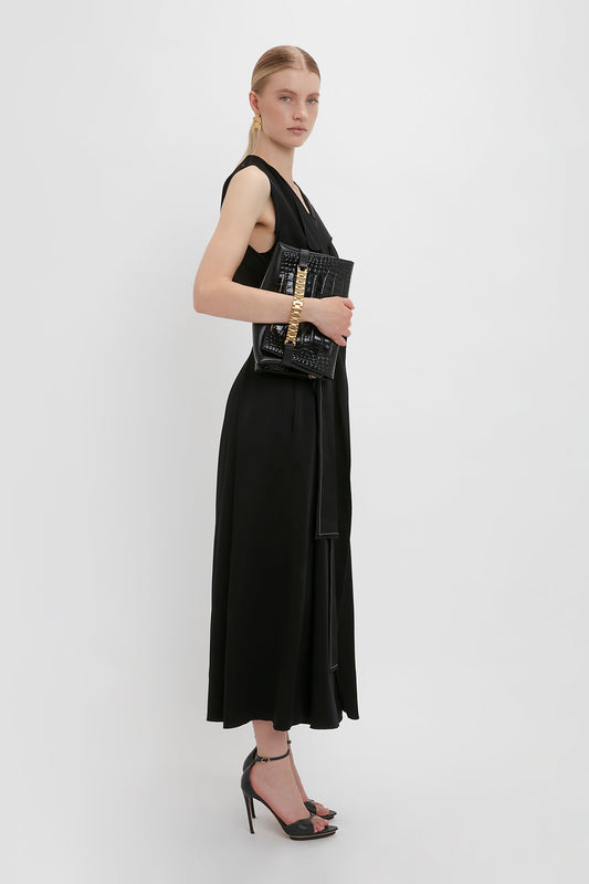 A person wearing a sleeveless Trench Dress In Black by Victoria Beckham and black high heels stands sideways, holding a black handbag with a gold chain.