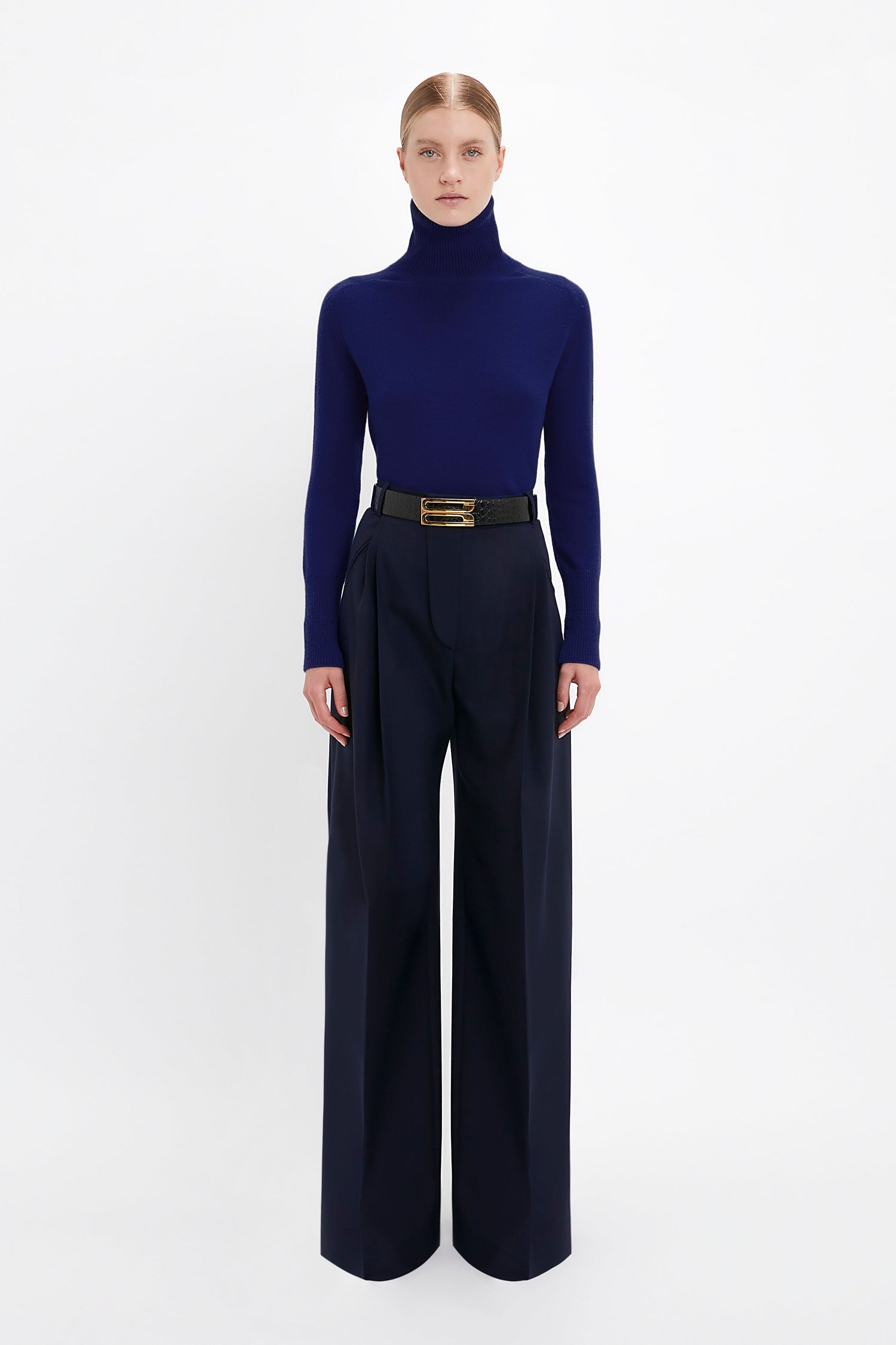 Person standing against a white background, wearing a dark blue Polo Neck Jumper In Navy by Victoria Beckham, black belt, and wide-leg navy trousers.