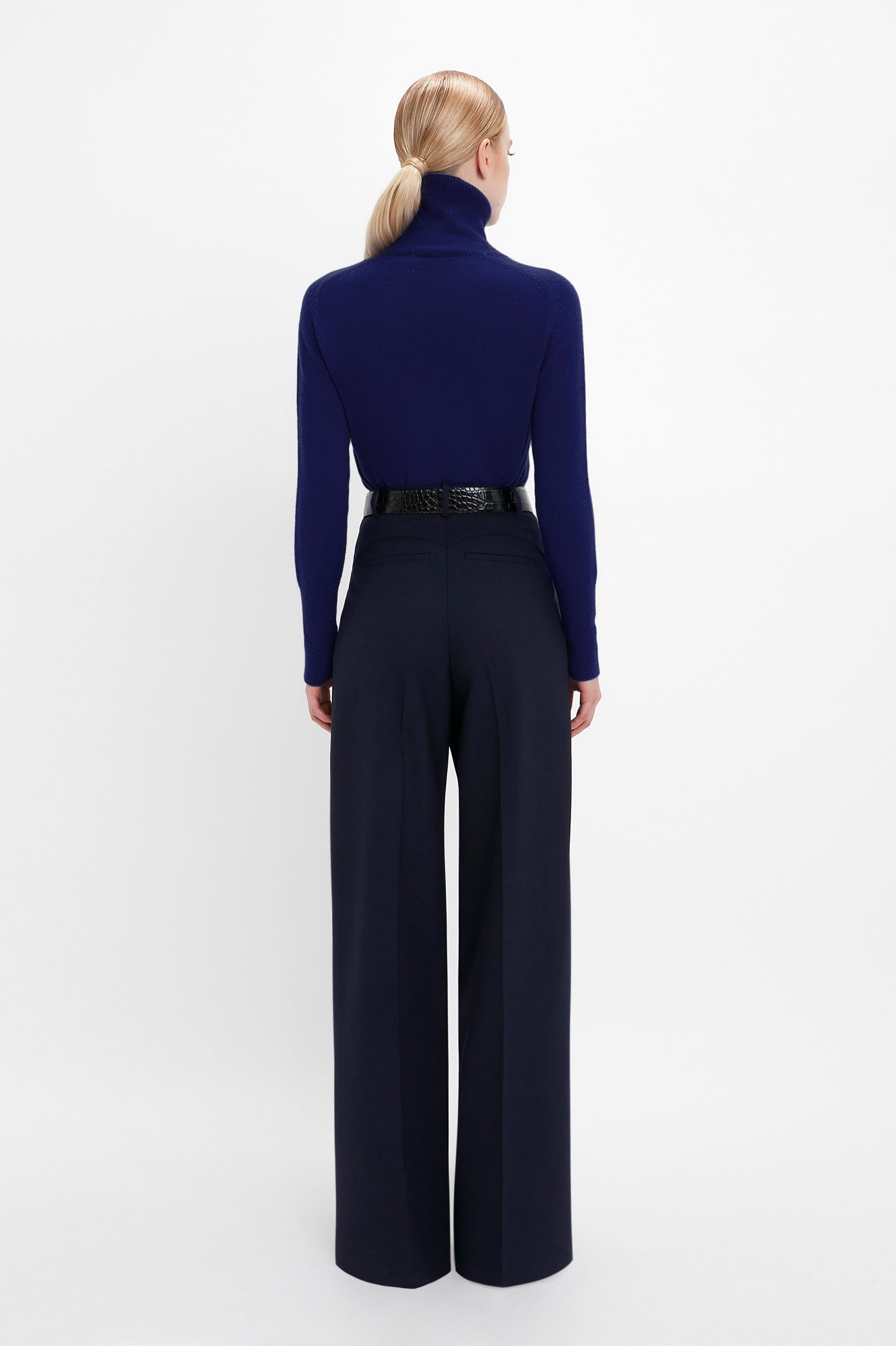 A person with blonde hair is seen from the back wearing a dark blue Polo Neck Jumper In Navy by Victoria Beckham tucked into high-waisted, wide-leg navy trousers with a black belt.