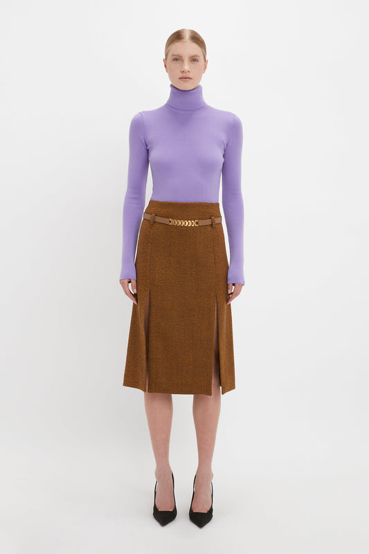A woman in a lilac turtleneck and brown skirt stands against a white background, wearing black Pointy Toe Pumps by Victoria Beckham.