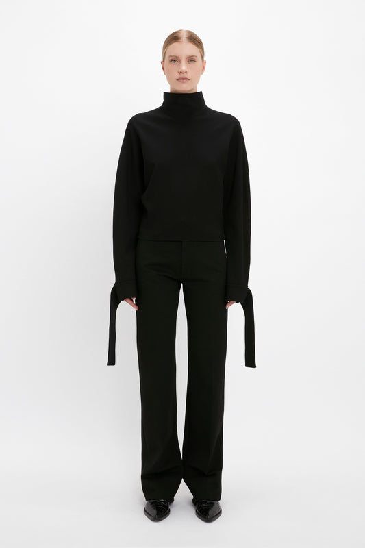 Person standing against a white background, dressed in an oversized slouchy fit black turtleneck "Tie Sleeve Ponti Top In Black" by Victoria Beckham with long sleeve extensions and black pants.