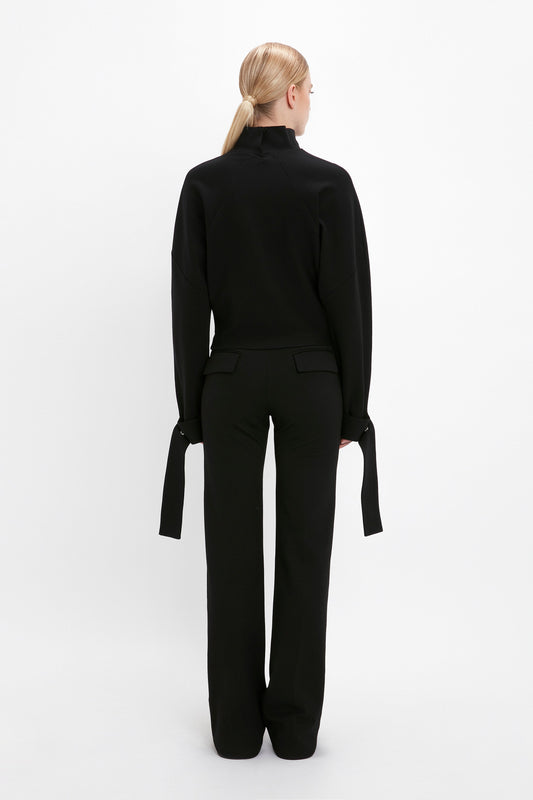 A person with light blonde hair stands facing away, wearing a black Tie Sleeve Ponti Top In Black by Victoria Beckham with long, loose cuffs and black pants against a plain white background.