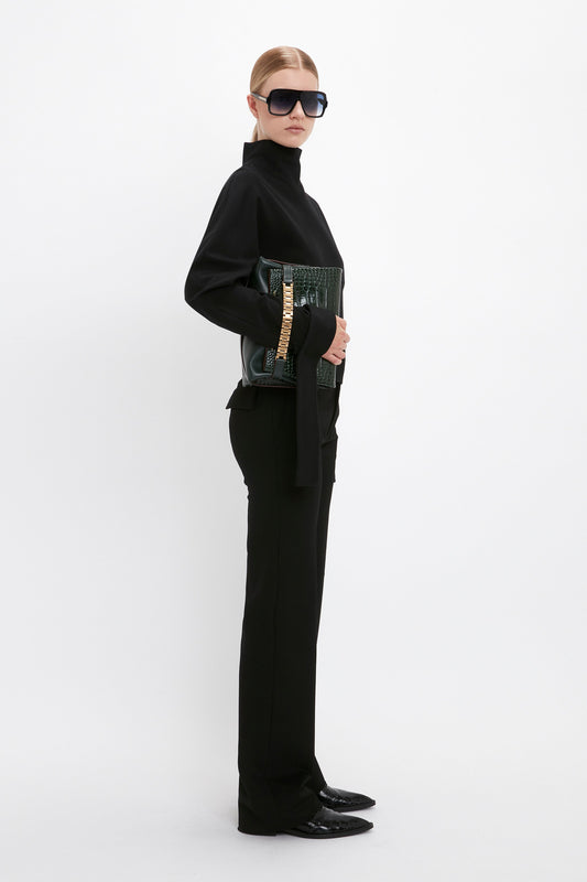 Person wearing a Victoria Beckham Tie Sleeve Ponti Top In Black, large dark sunglasses, and holding a dark green handbag with a gold chain detail, stands against a plain white background.
