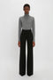 A person stands against a plain white background wearing a Victoria Beckham Polo Neck Jumper In Grey Melange and black high-waisted, wide-leg pants, exemplifying versatile styling.