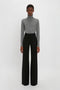 A person stands against a plain background, wearing a gray turtleneck sweater and black high-waisted Alina Tailored Trouser In Black by Victoria Beckham, exuding a timeless 70s style reminiscent of Victoria Beckham's elegant fashion sense.