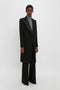 Person standing against a white background wearing a long black Double Breasted Tuxedo Coat in Black by Victoria Beckham over a grey turtleneck and black wide-leg pants, exuding a modern relaxed feel with the lightweight wool gabardine fabric.