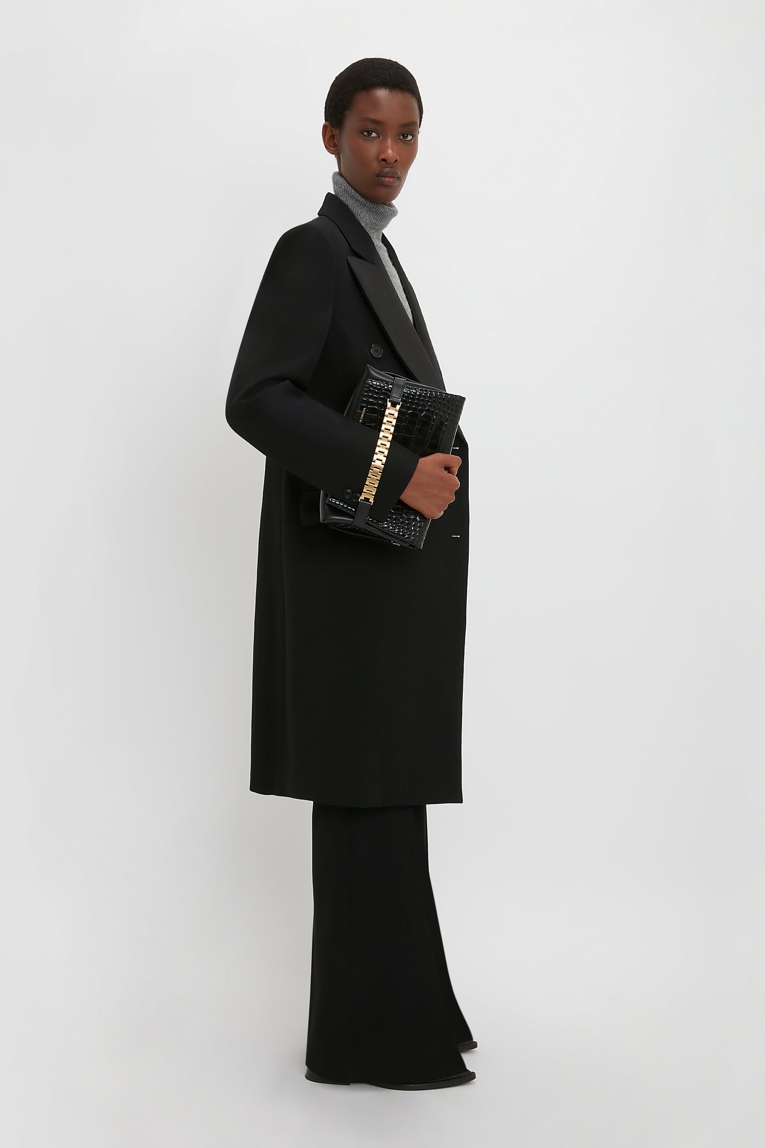 A person wearing a Victoria Beckham Double Breasted Tuxedo Coat in Black, black flared trousers, and a grey turtleneck sweater holds a textured black clutch against a plain white background. The ensemble captures a modern relaxed feel with its lightweight wool gabardine fabric.