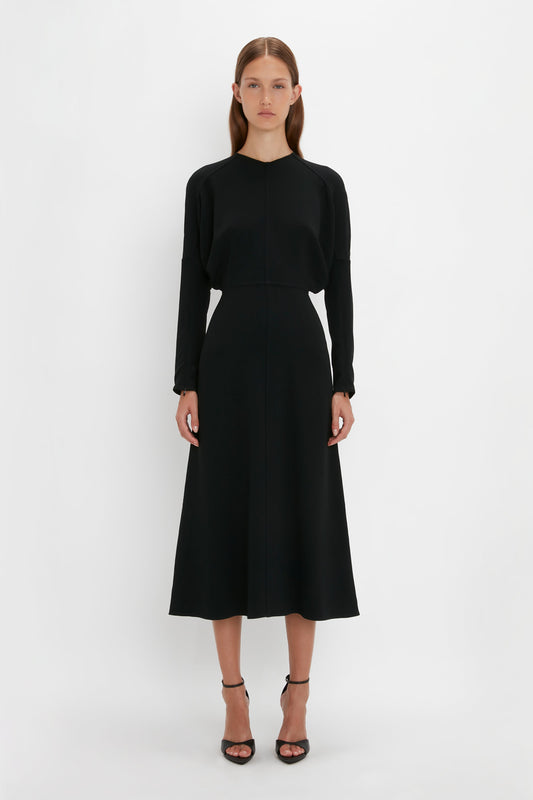 A woman in a plain, elegant black Victoria Beckham dolman sleeve midi dress with long sleeves and pointy toe stiletto sandals, standing against a white background.
