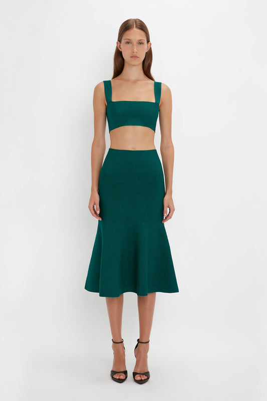 A woman stands against a plain background, wearing a VB Body Strap Bandeau Top In Lurex Green and matching VB Body flared skirt. She has straight hair and is wearing black pointy toe stiletto sandals.