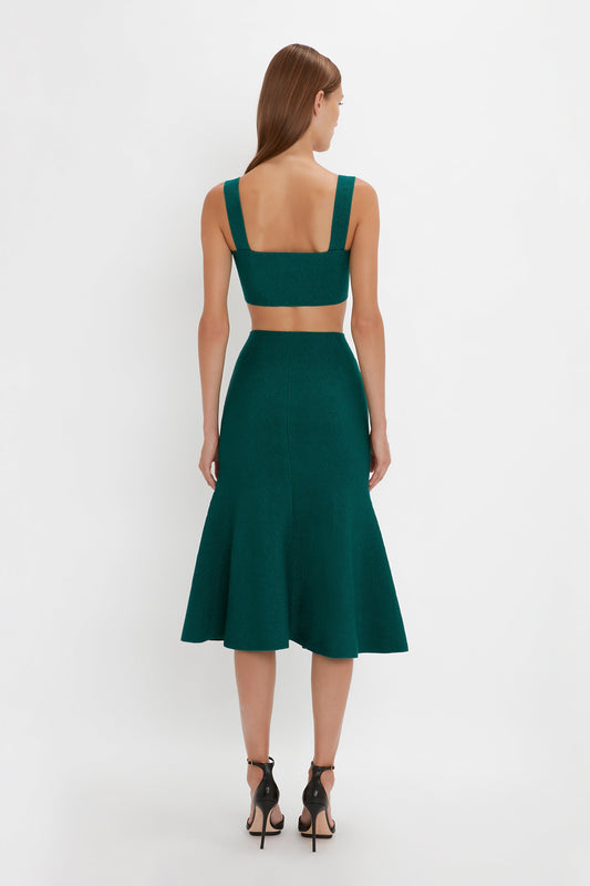 A woman with long hair wears a dark green, backless, sleeveless dress with a flared silhouette and black high-heeled shoes from Victoria Beckham, specifically the VB Body Flared Skirt In Lurex Green, standing and facing away from the camera against a plain white background.